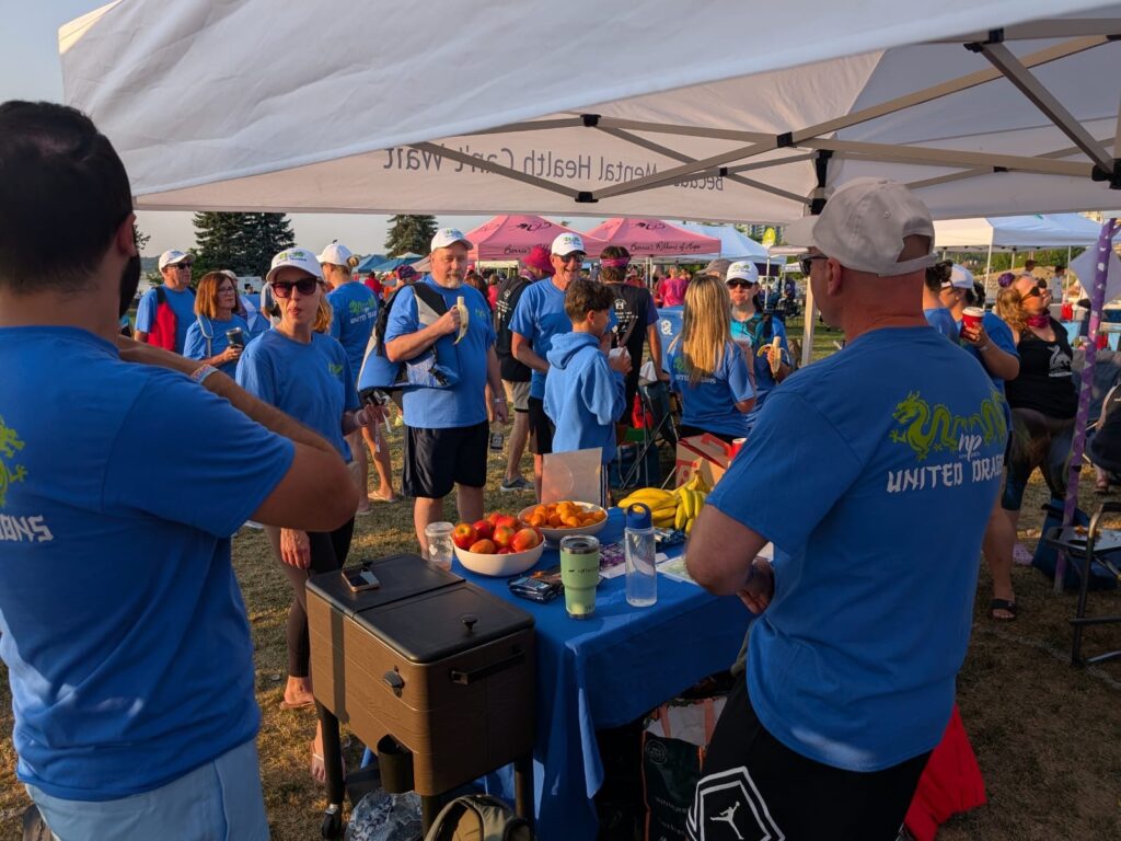 Group of people wearing matching white hats and blue New Path United Dragons t-shirts standing under a tent in front of and behind a table with food on it.