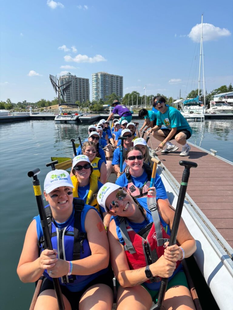Group of people wearing life jackets and matching white hats holding paddles in a dragon boat and smiling.