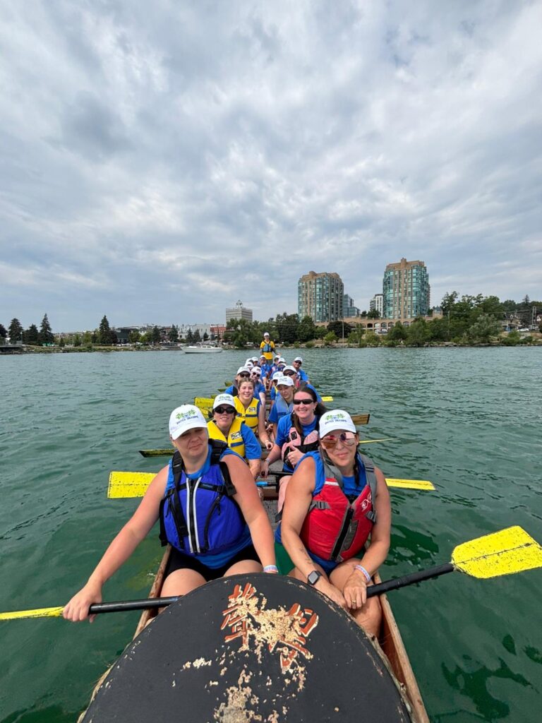 Group of people wearing life jackets and matching white hats holding paddles in a dragon boat and smiling.