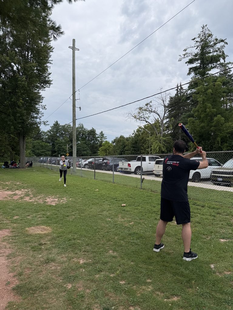 One person holding a bat and another one pitching to them in a park with cars parked to the side.