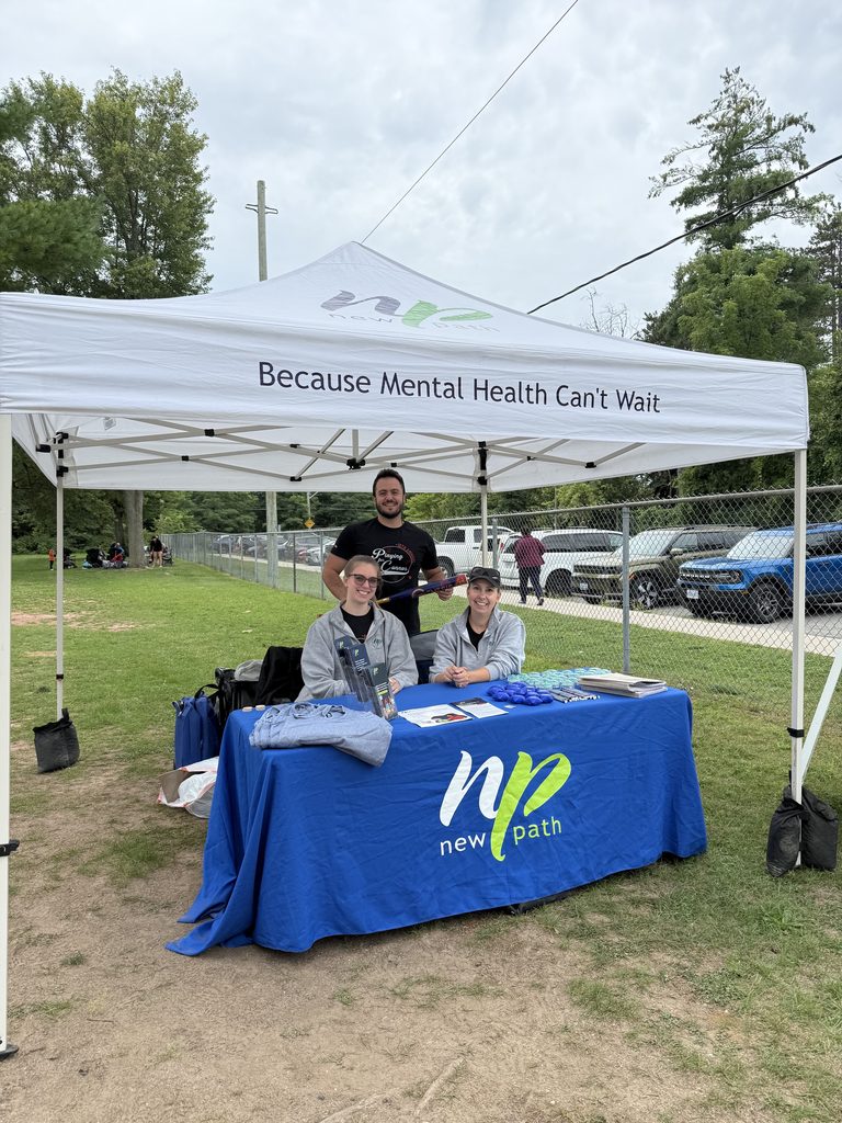 Picture of three people behind a New Path branded table cloth and under a tent that says Because Mental Health Can't Wait. 