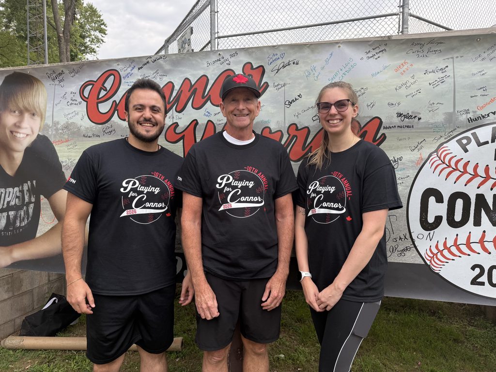 Three people wearing Playing for Connor t-shirts smiling with a Playing for Connor banner in the background. 
