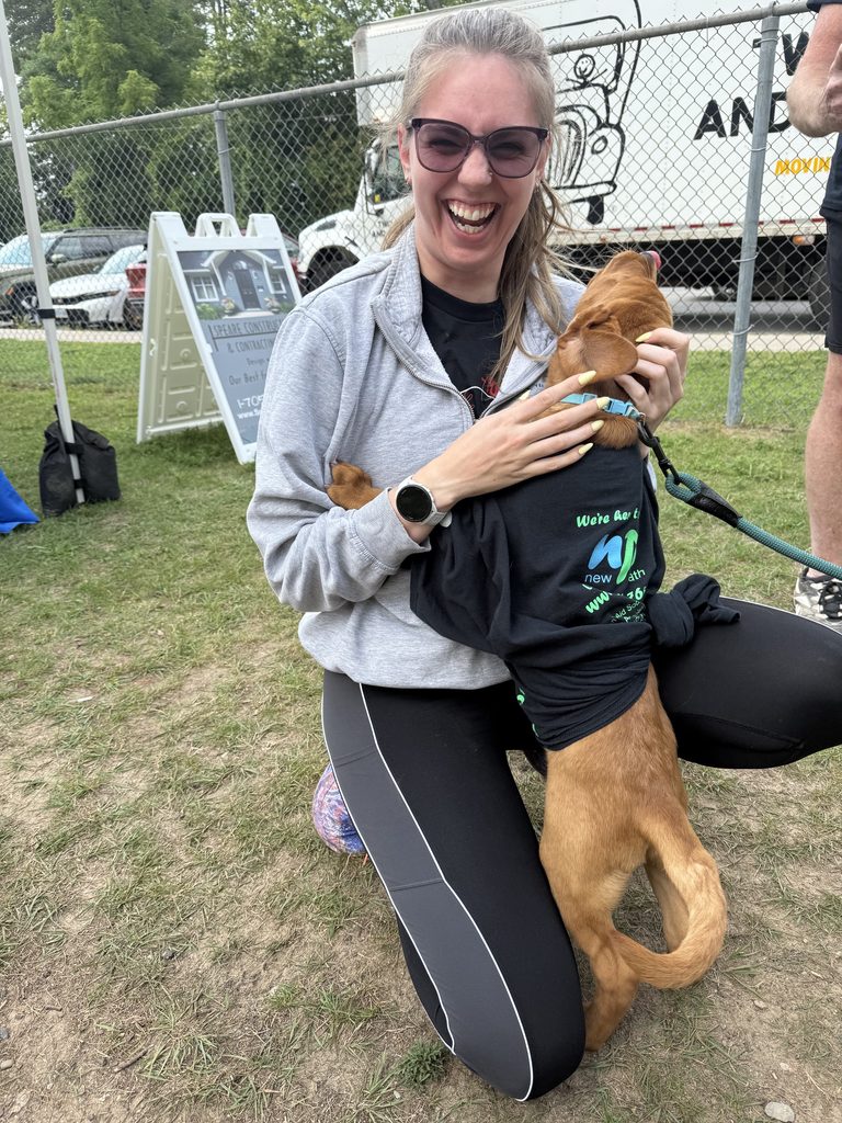 A woman laughing while a small dog jumps up on her. The dog is wearing a New Path t-shirt. 