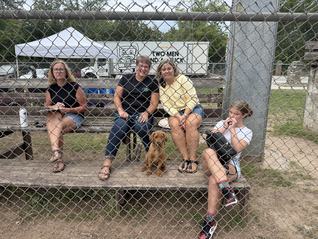 Picture of three women and a dog sitting on bleachers behind a fence. 