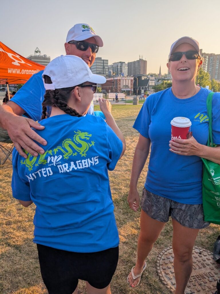 Picture of three people smiling and wearing matching New Path United Dragons t-shirts. Two of the three are embracing in a hug.